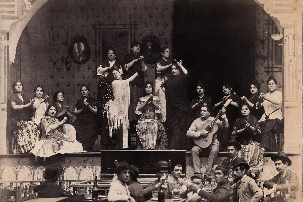 Čiernobiela fotografia café cantante v Seville okolo roku 1888, s dvoma ženami tancujúcimi a ostatnými tlieskajúcimi na vyvýšenom pódiu, gitarista sediaci vpravo a muži pri stoloch vpredu