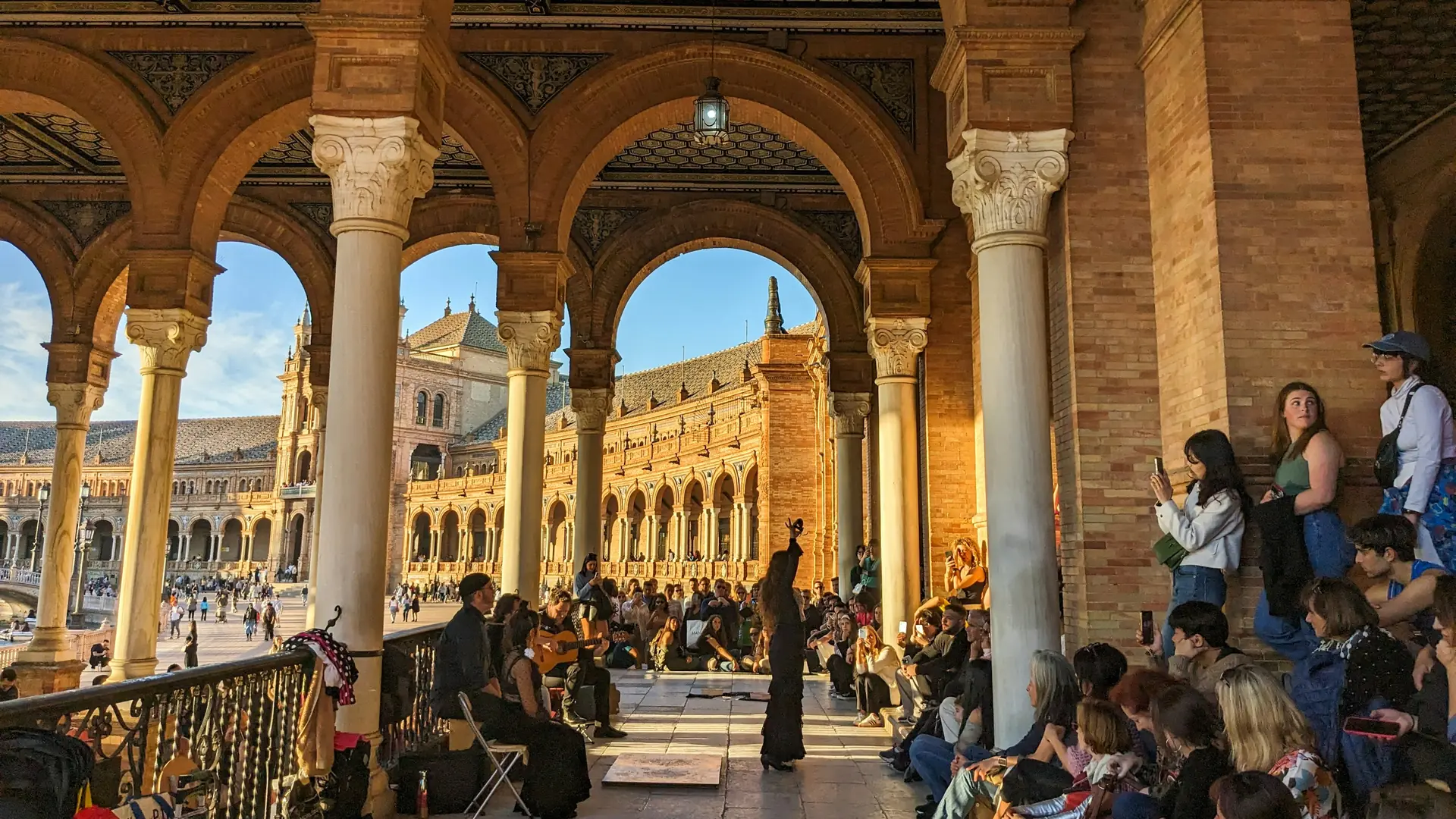 Flamencová tanečnice v černém zvedá jednu paži ve zlatém světle pod cihlovými arkádami Plaza de España v Seville, vedle ní kytarista a kolem ní sedící publikum